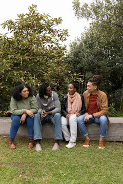Diverse friends sitting on bench in park chatting, holding phone, wearing scarves and jeans