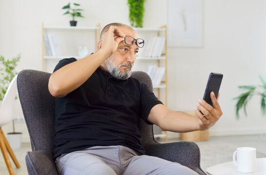 Senior man checking smartphone trying to read message having farsightedness eye disease. At home on chair, he wears glasses, focused on screen in living room. Presbyopia and vision problems concept.