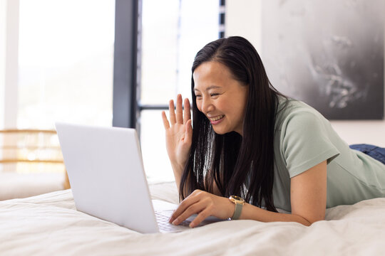 Asian woman lying on white duvet, propping on elbows, wearing light green shirt, waving at laptop