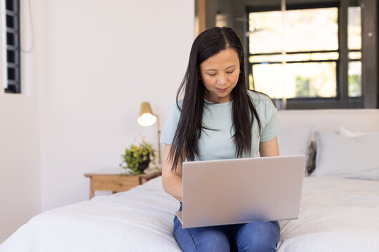 Asian woman sitting on neatly made bed using silver laptop in softly lit bedroom with lamp