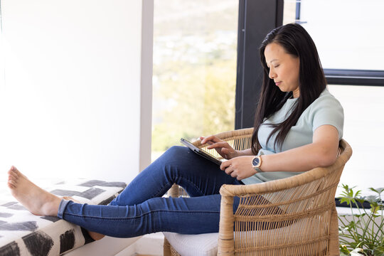 Asian woman sitting in wicker armchair tapping tablet on patterned ottoman at window, copy space