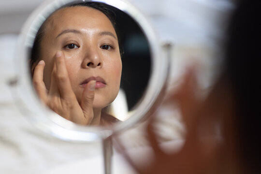 Asian woman inspecting face in round magnifying mirror at vanity, applying makeup with fingertips