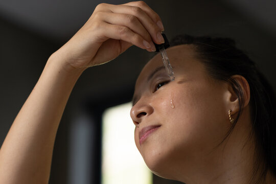 Adult Asian woman applying clear serum with glass dropper near eye in softly lit room