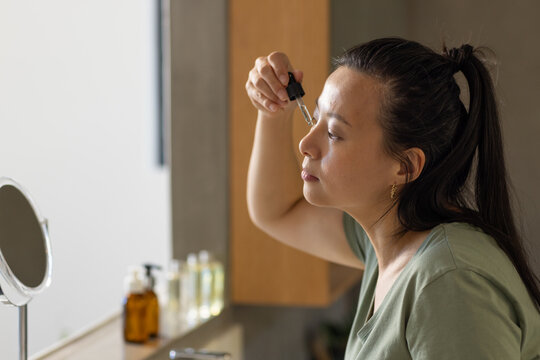 Asian woman applying serum with glass dropper at vanity near mirror and amber bottles