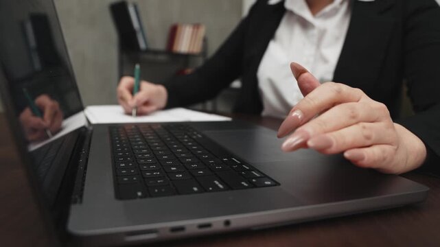A businesswoman types on a laptop while taking notes on paper in a modern office interior with shelves and books in the background.