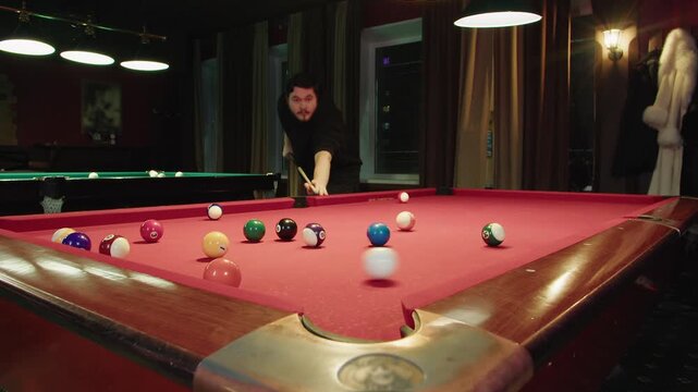 Wide angle view of caucasian young man that plays billiards and hits the white ball with a cue. Nightlife ambiance and intense pool game in a club. The ball rolls on the red table and hits the sides