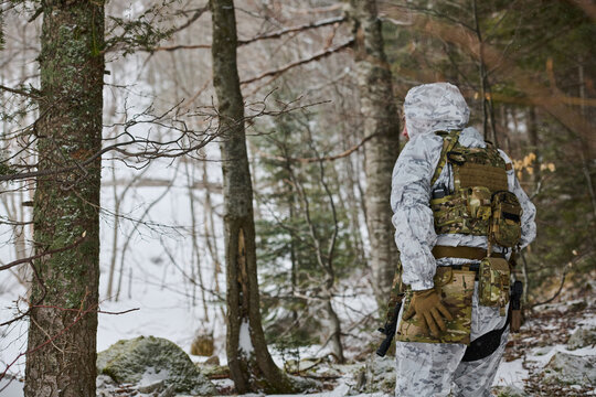 Soldier In White Winter Camouflage Patrols Snowy Forest With Tactical Gear, Backpack And Rifle
