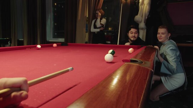 Group of joyful friends enthusiastically watches cue ball contact on red table. Young caucasian man and a woman show upset emotions after missing a shot. Social interaction and leisure in a club