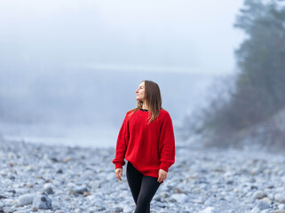 Young woman in a vibrant red sweater stands on a rocky riverbank, surrounded by misty landscape, embodying tranquility and connection with nature in a serene environment © Ktn