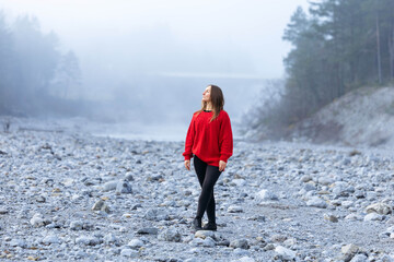 Woman in a red sweater stands on a rocky riverbed, surrounded by misty landscape, showcasing a serene moment in nature with a tranquil atmosphere and soft light © Ktn