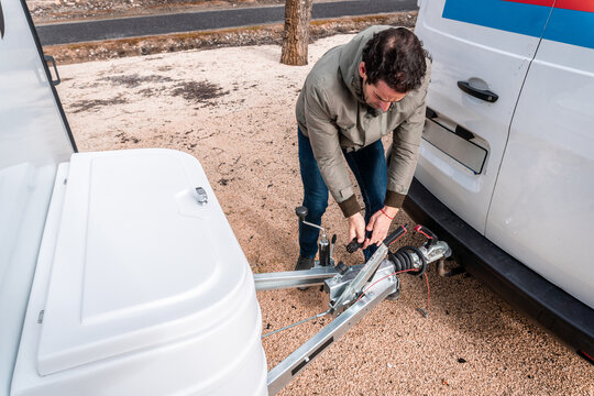 Man hooks small cargo trailer to white van at gravel roadside, preparing for travel, delivery or camping trip with safety checks