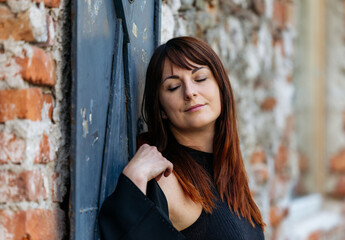 Fototapeta premium Woman with long brown hair stands against a rustic brick wall, eyes closed, exuding a sense of calm and tranquility in a serene urban environment