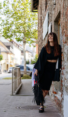 Stylish woman in black outfit with fringe bag stands against a rustic stone wall, smiling and enjoying a sunny day in an urban environment, showcasing modern fashion and confidence © Ktn