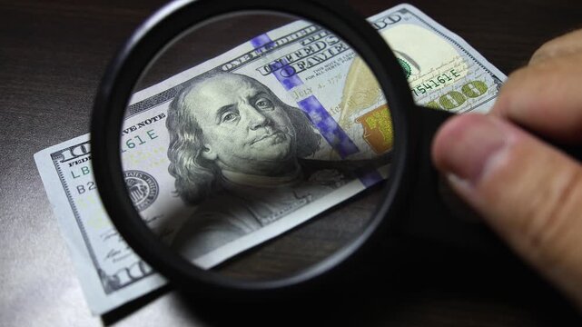 Examination of american dollar (USD) cash money with magnfication glass, loupe. Concept of authenticity check, fight against counterfeiting of banknotes, anti fraud measures in USA. Close up view.
