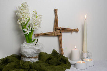 A Saint Brigid’s Imbolc altar with a woven Brigid’s cross, white hyacinth, lit candles, and green cloth is arranged against a light background to symbolize purity, renewal, and early spring.