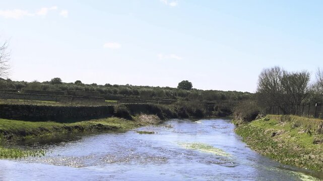 Video from a bridge overlooking a calm river curving through green banks, with ripples, reflections and patches of algae under bright blue sky.