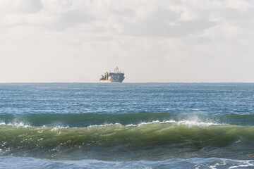 Fototapeta premium A large cargo ship sits on the calm horizon of a blue ocean, with a gentle green wave breaking in the foreground under a soft, hazy sky. A peaceful maritime scene