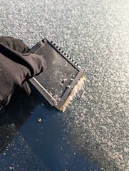 Close up of a gloved hand using a black plastic scraper to remove thick white frost from a car windshield
