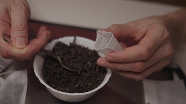 Close-up top view of hands preparing individual tea bags at home, scooping loose leaf black tea from white bowl into small paper packet for daily brewing, closeup, slow motion.