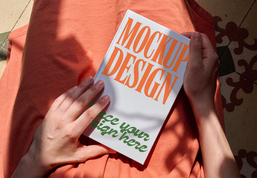 A blank notebook cover held by woman in an orange outfit, illuminated by soft sunlight casting shadows in a cozy setting. PSD template mockup.