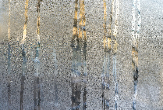 Condensation covering wet glass surface with flowing water streaks. moisture creating abstract pattern and blurred light shapes, closeup background texture, humidity concept, cold weather atmosphere,