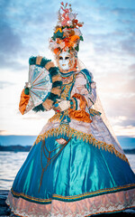 Obraz premium Venice, Italy - February 09, 2026: Elegant masked figure in ornate costume holds a fan at the Venice Carnival, showcasing intricate details and vibrant colors against a scenic backdrop