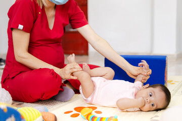 A professional woman performs stimulation and child care exercises with a 6-month-old baby during a session in a clinic, a pregnant healthcare worker wearing red fluid-resistant clothing.