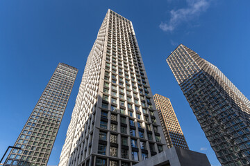 Obraz premium Tall buildings stand in a row against a blue sky. The scene shows a modern city atmosphere with glass and concrete structures reflecting sunlight. People walk below, enjoying the day.
