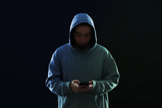 Criminal in hoodie using mobile phone on black background. Man with unrecognizable pixelated face