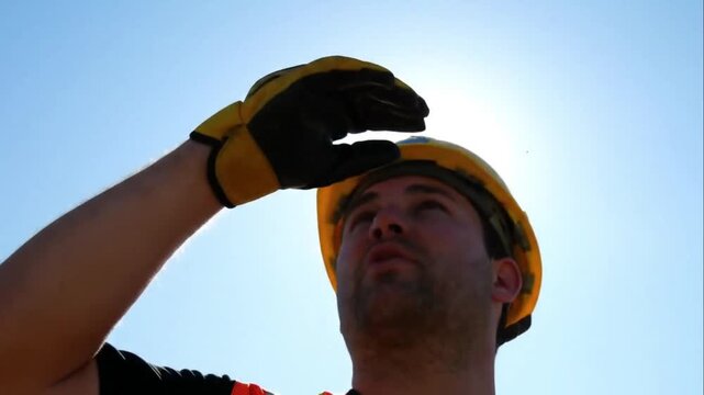 Low angle, A construction worker wiping sweat from their forehead with a gloved hand, sun flaring behind them.