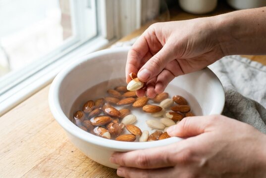 Woman's hands peeling the skin from a soaked almond over a white bowl of water, preparing for cooking or making milk.