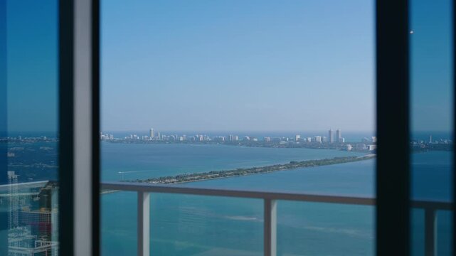 Coastal view shows calm water stretching across scene, bridge connects land, distant skyline rises with tall buildings, balcony railing and window frame add depth and clarity
