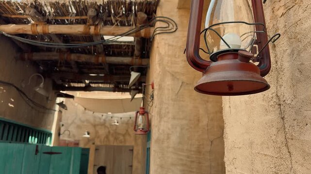 Close-up of a rustic brown lantern hanging on a textured wall in a traditional Arab heritage village, featuring wooden ceiling beams and a narrow alleyway in the background.

