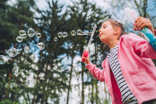 Young girl in pink jacket and striped shirt blowing soap bubbles with plastic wand in green park during summer featuring blurry pine trees background area and magical flying bubbles in sunlight