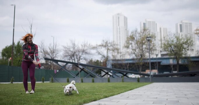 Woman walking white dog on leash in City Park during daytime hours wearing athletic clothes, the dog scratches an itch on its body