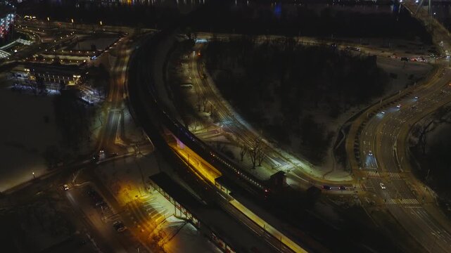 Aerial night over Warsaw, Poland shows Vistula River, PGE Narodowy, Swietokrzyski Bridge, snow dusted streets, amber lit station, moving cars and a passing train.