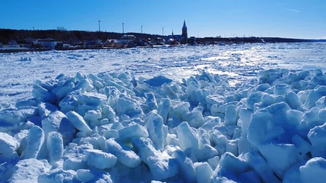 A frozen coastal landscape, made of cracked pieces of ice and bathed in sunlight, with a village and church steeple along the shore and wind turbines visible on the distant horizon. Baie-des-Sables.