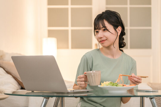 Young Asian woman relaxing and looking at laptop while eating at home in the evening