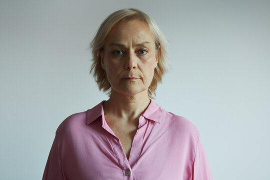Portrait of senior serious woman in pink shirt looking at camera, natural light, neutral background