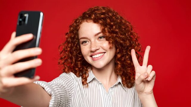 A young woman with curly red hair and freckles takes a selfie with her phone, making a peace sign and puckering her lips against a vibrant red background. She wears a striped shirt.