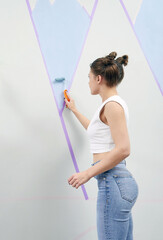 Young woman painting wall with paint roller and using masking tape while standing on ladder