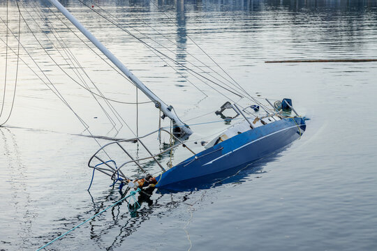 Sunken blue sailboat partially submerged in calm water with tilted hull, mast, and rigging visible creating reflections
