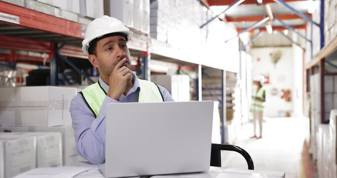 Serious young Hispanic warehouseman in hard hat making inventory, counting boxes on metal shelves after shipment, consulting paper waybill and laptop computer, working in industrial storage