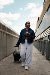 Young black woman walking with suitcase and using smartphone near train station