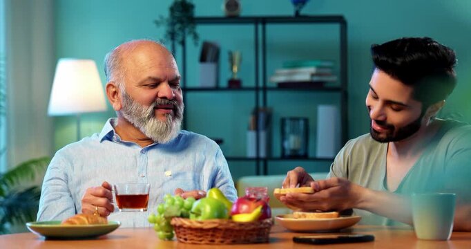 Indian young son chatting with senior father while having breakfast together in modern home, happy morning family bonding over bread jam coffee, healthy lifestyle conversation between two generations