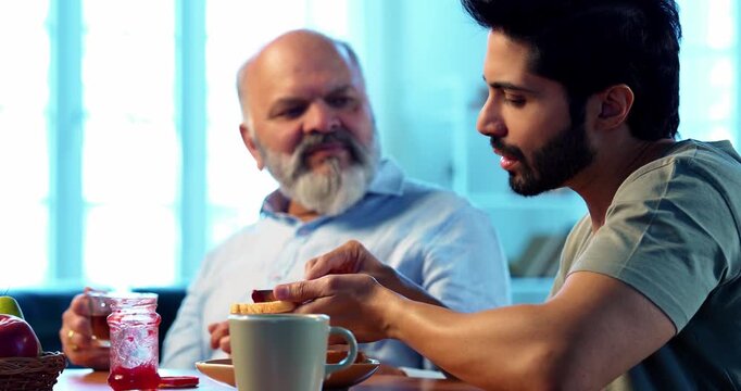Indian young son chatting with senior father while having breakfast together in modern home, happy morning family bonding over bread jam coffee, healthy lifestyle conversation between two generations