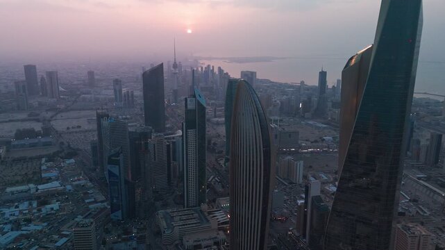 High-angle aerial at sunset over Kuwait City, showing dense skyscrapers, landmark towers, and the coastline beside the Arabian Gulf in warm haze and soft evening light.