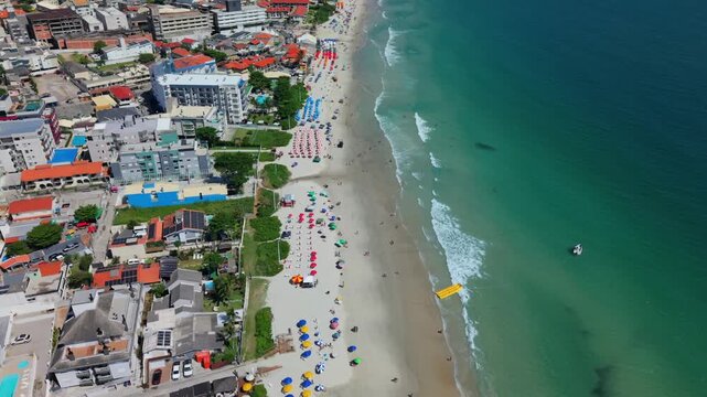 Downward tilted aerial glide over turquoise waters and beach umbrellas. The clip moves toward a residential area at Praia do Santinho in Florian&oacute;polis, Santa Catarina, Brazil.
