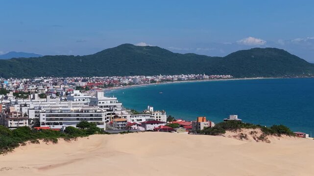 Forward aerial glide over white sand dunes towards a calm bay and residential area. Green hills surround the landscape at Praia do Santinho in Florian&oacute;polis during a sunny day.