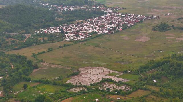 Aerial drone footage over rural village and rice fields showing patchwork paddies irrigation ponds tropical green hills forest edge and wide farmland valley countryside landscape in soft haze 4k